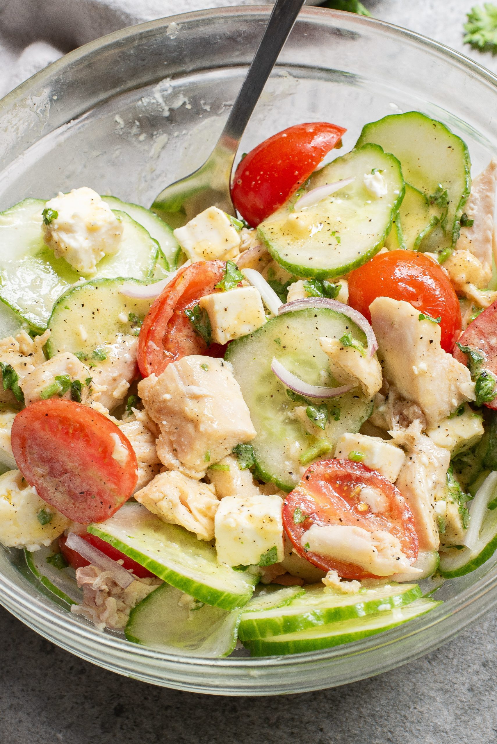 A clear bowl filled with a salad of cucumber slices, cherry tomatoes, diced chicken, feta cheese, and chopped herbs, with a fork.