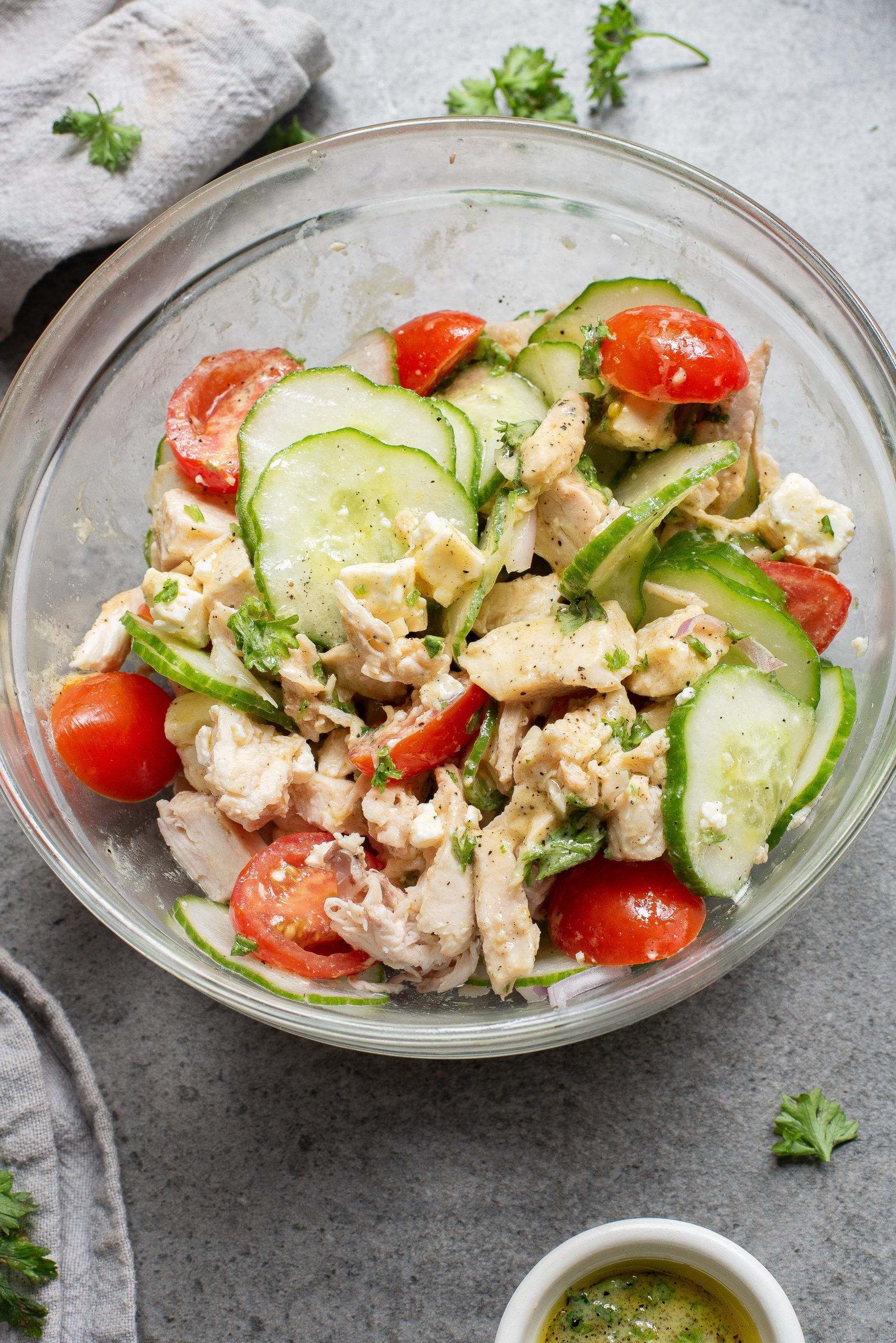 Glass bowl with a chicken salad featuring sliced cucumbers, cherry tomatoes, and parsley. A small bowl of dressing is visible beside it.