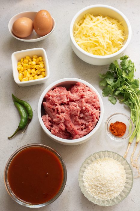 Ingredients on a surface: ground meat, grated cheese, two eggs, corn, coriander, green chilies, breadcrumbs, red spice, and sauce in separate containers.