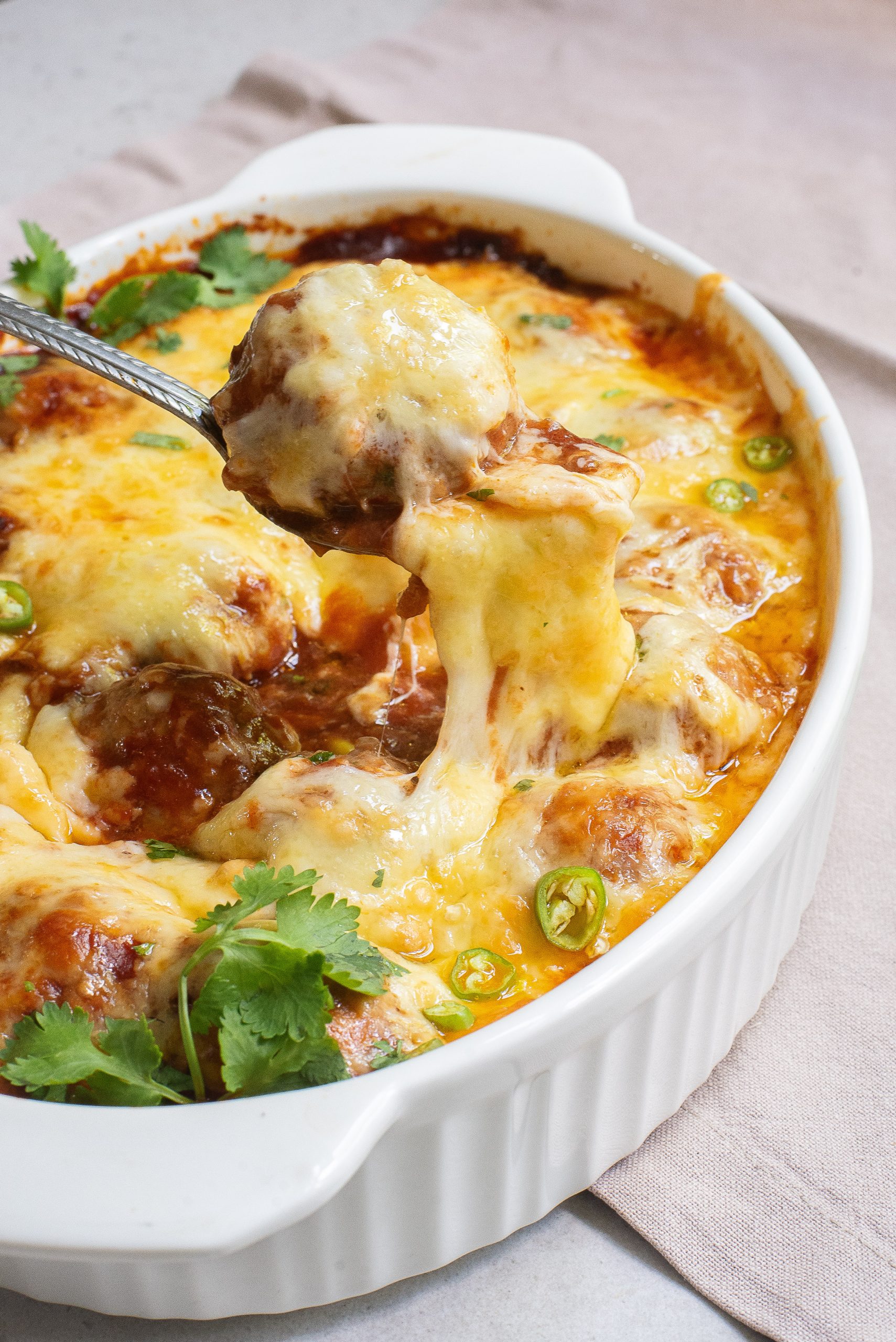 A white dish filled with baked meatballs covered in melted cheese, garnished with sliced green peppers and fresh cilantro. A spoon lifts a meatball, showing cheese stretching.