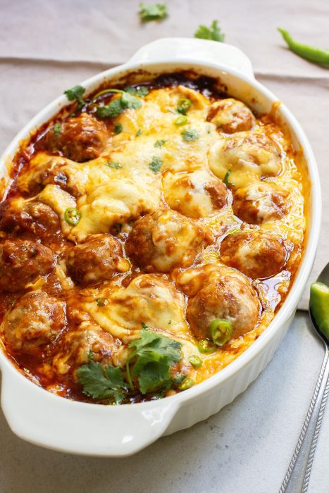 Baked meatballs in a white dish, covered with melted cheese and garnished with cilantro, on a light-colored surface.