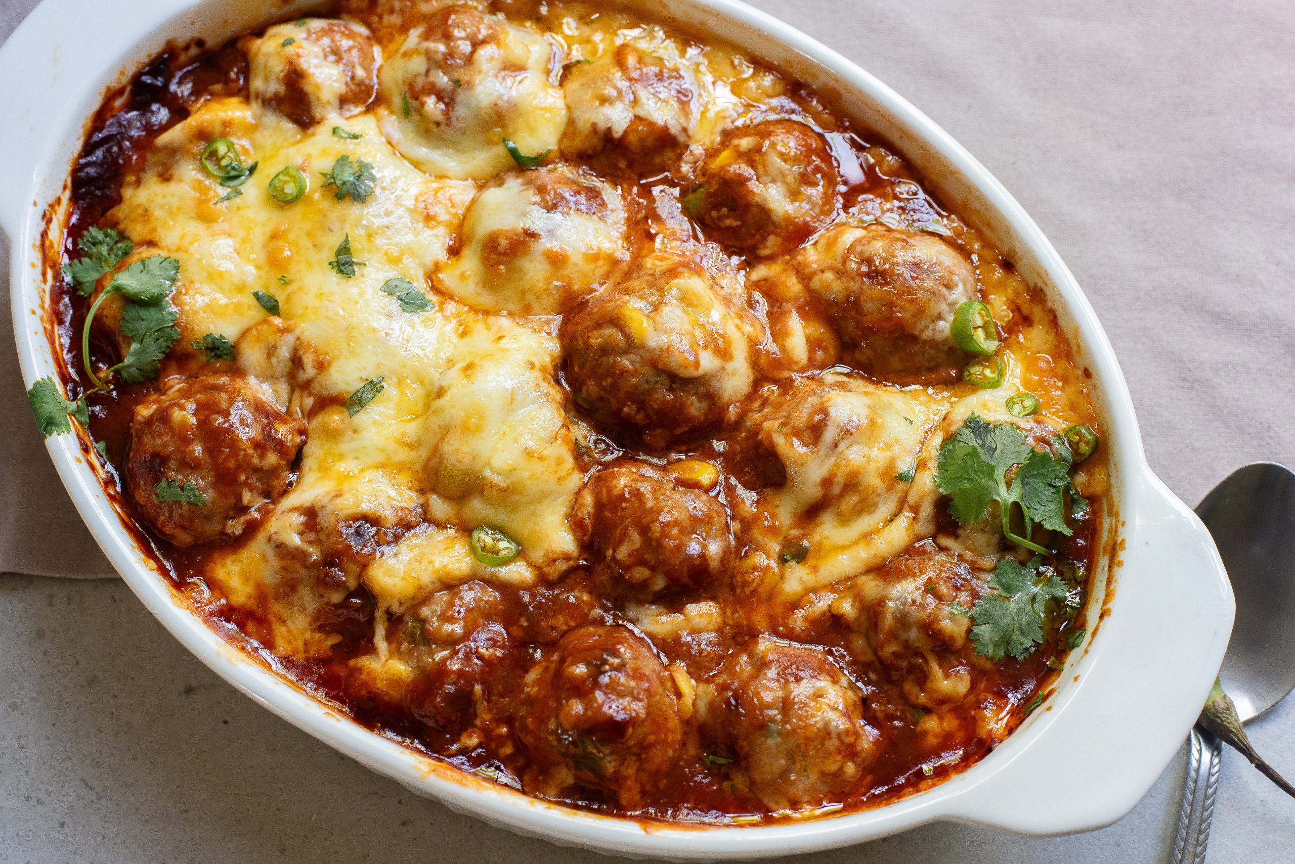 A baked dish with meatballs topped with melted cheese and garnished with cilantro in an oval ceramic casserole.