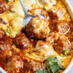 A dish of baked meatballs in a rich tomato sauce topped with melted cheese, garnished with cilantro, with a spoon resting on top.