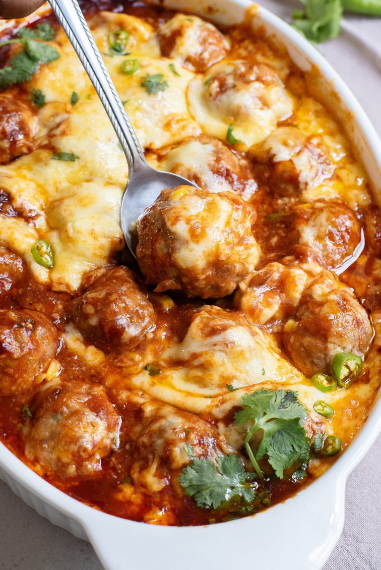 A dish of baked meatballs in a rich tomato sauce topped with melted cheese, garnished with cilantro, with a spoon resting on top.