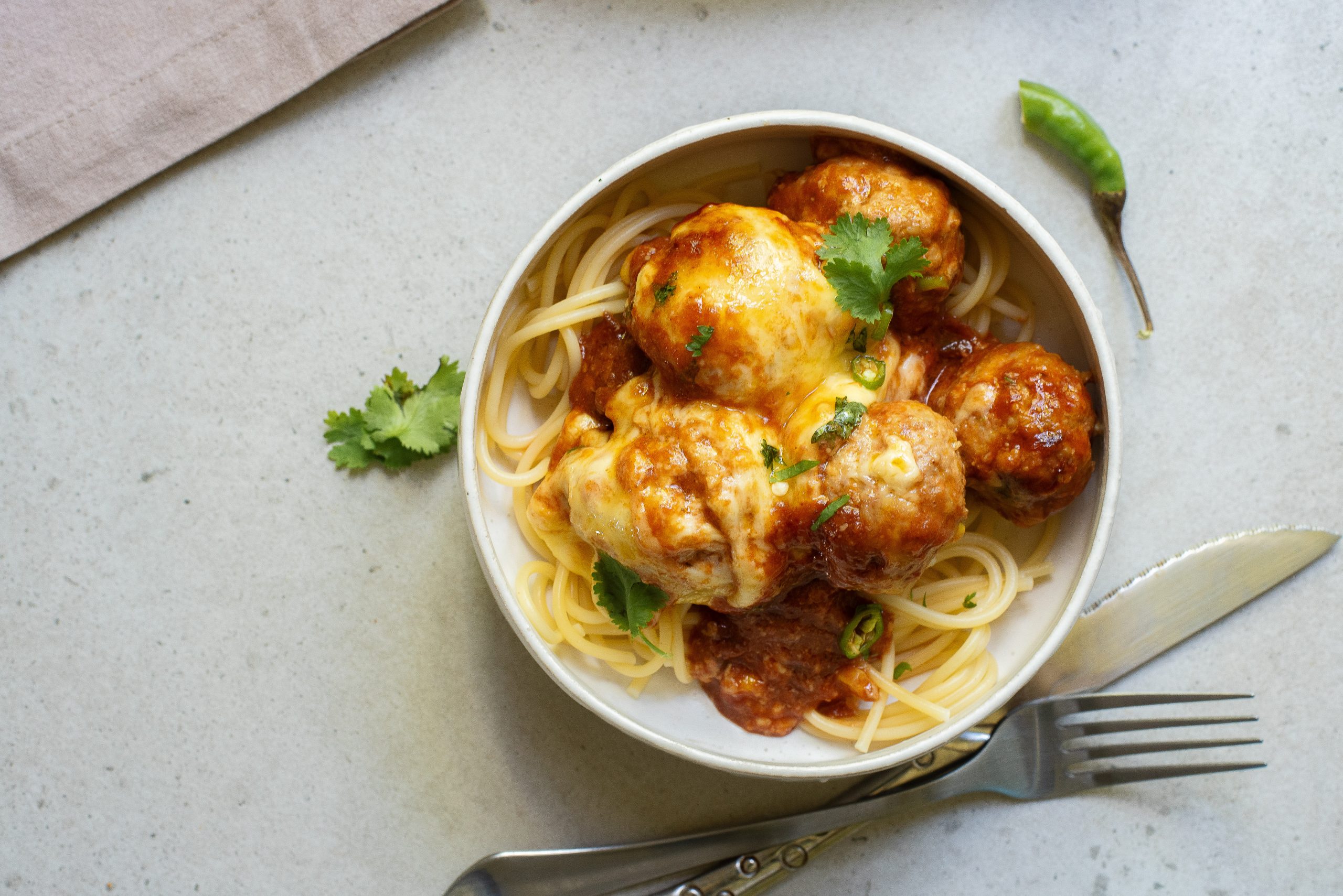 Bowl of spaghetti topped with meatballs, tomato sauce, melted cheese, and garnished with cilantro. A fork and knife are placed beside the bowl on a light surface.