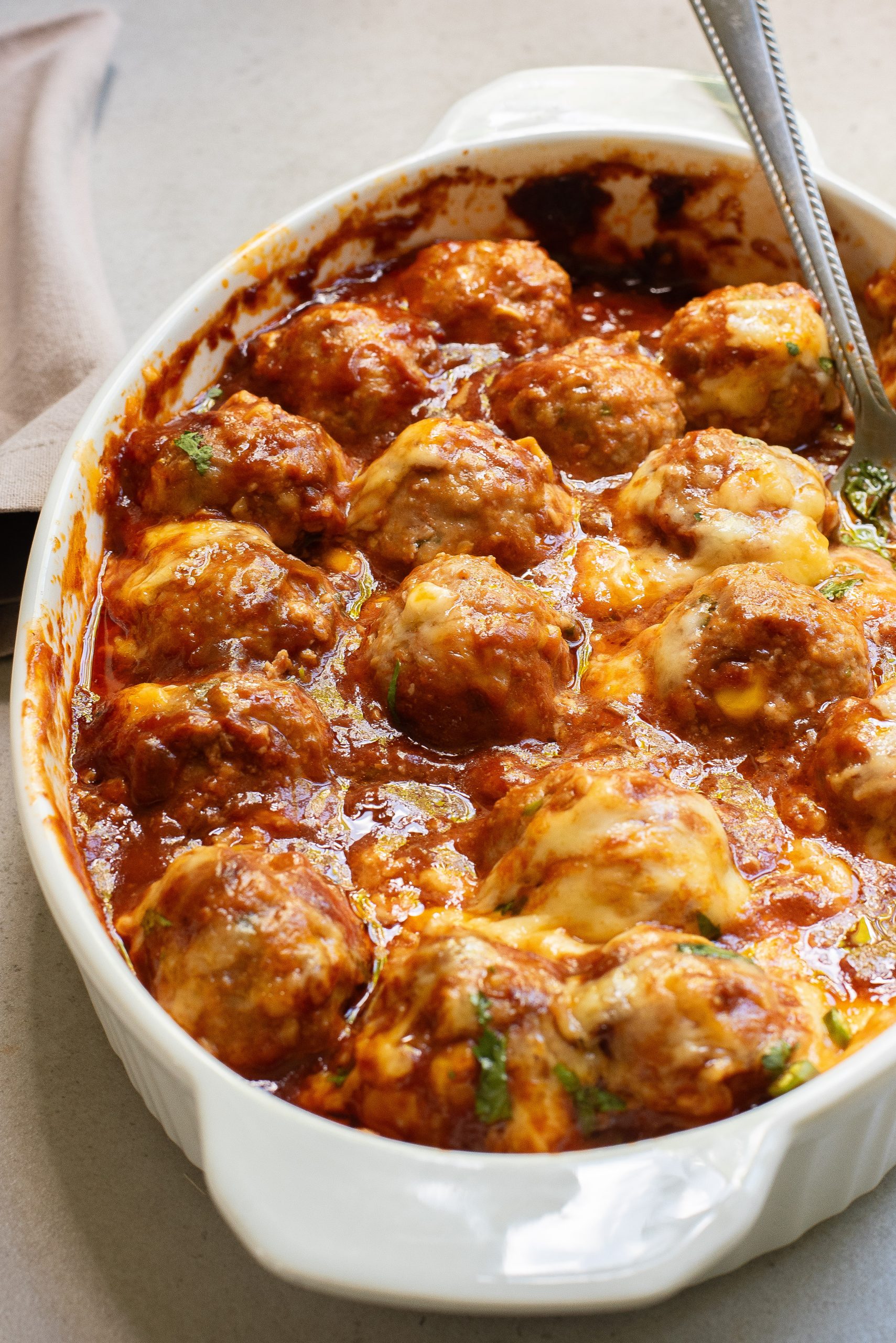 A white dish filled with baked meatballs covered in tomato sauce and melted cheese, with a spoon resting inside.