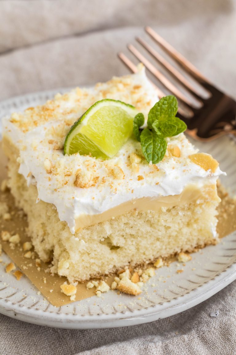 A slice of cake topped with whipped cream, crumbled nuts, a lime wedge, and a mint sprig on a white plate. Two copper forks are placed beside the plate.