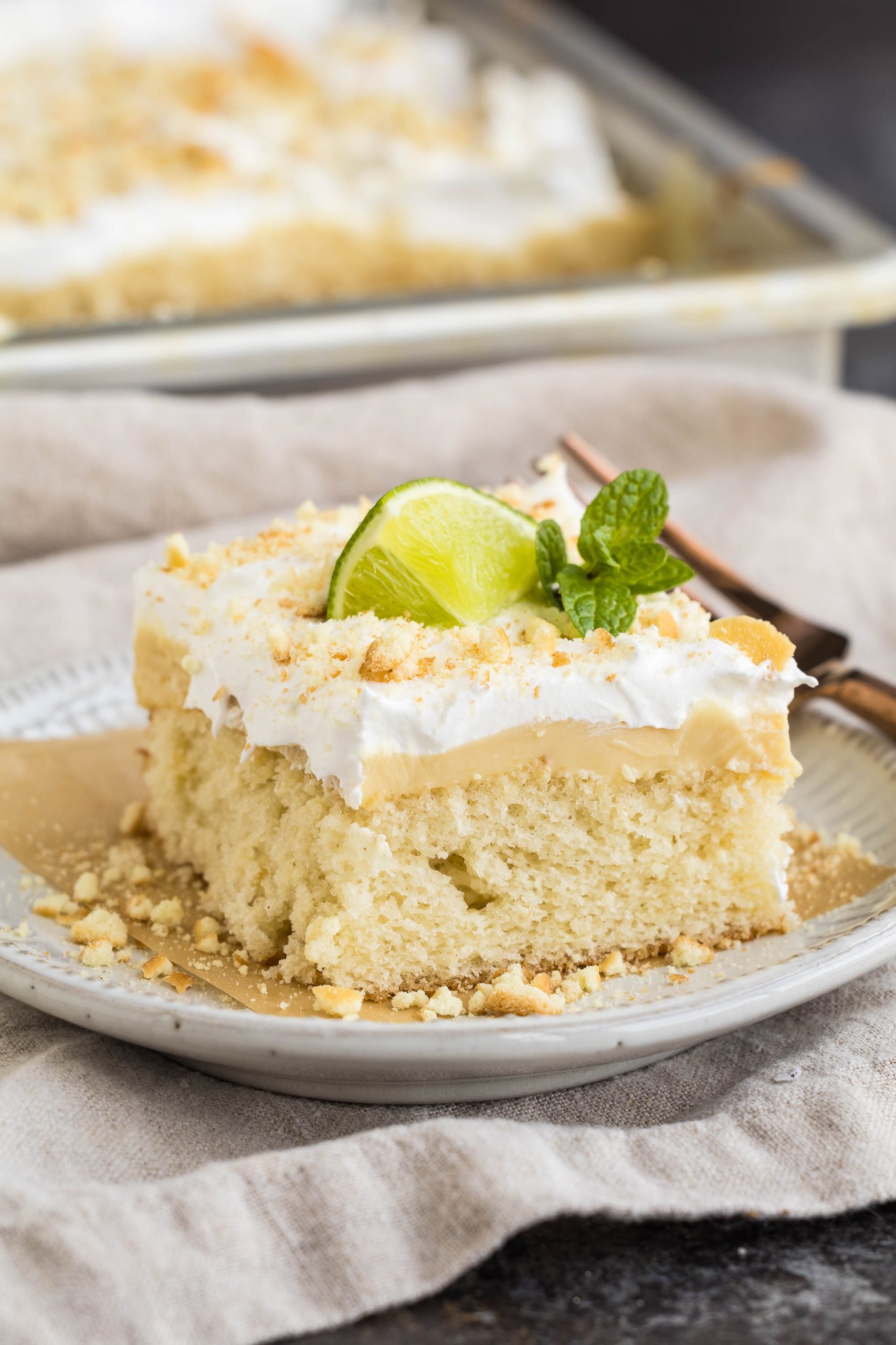 A slice of Key Lime Pie Sheet Cake with creamy frosting and crumbled topping, garnished with a lime wedge and mint leaf, served on a plate.