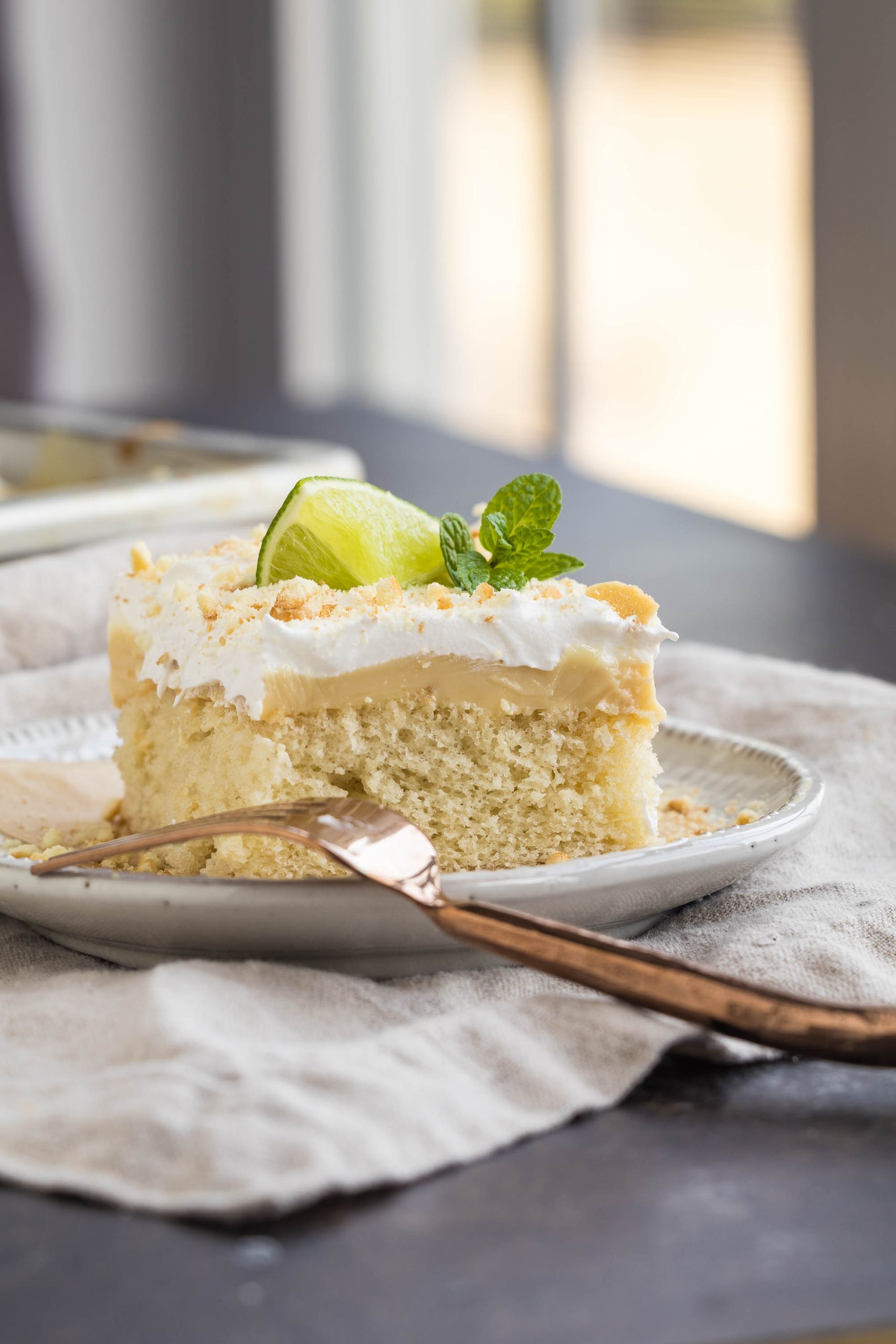 A slice of cake with white frosting, garnished with a lime wedge and mint, is served on a plate with a fork on a cloth.