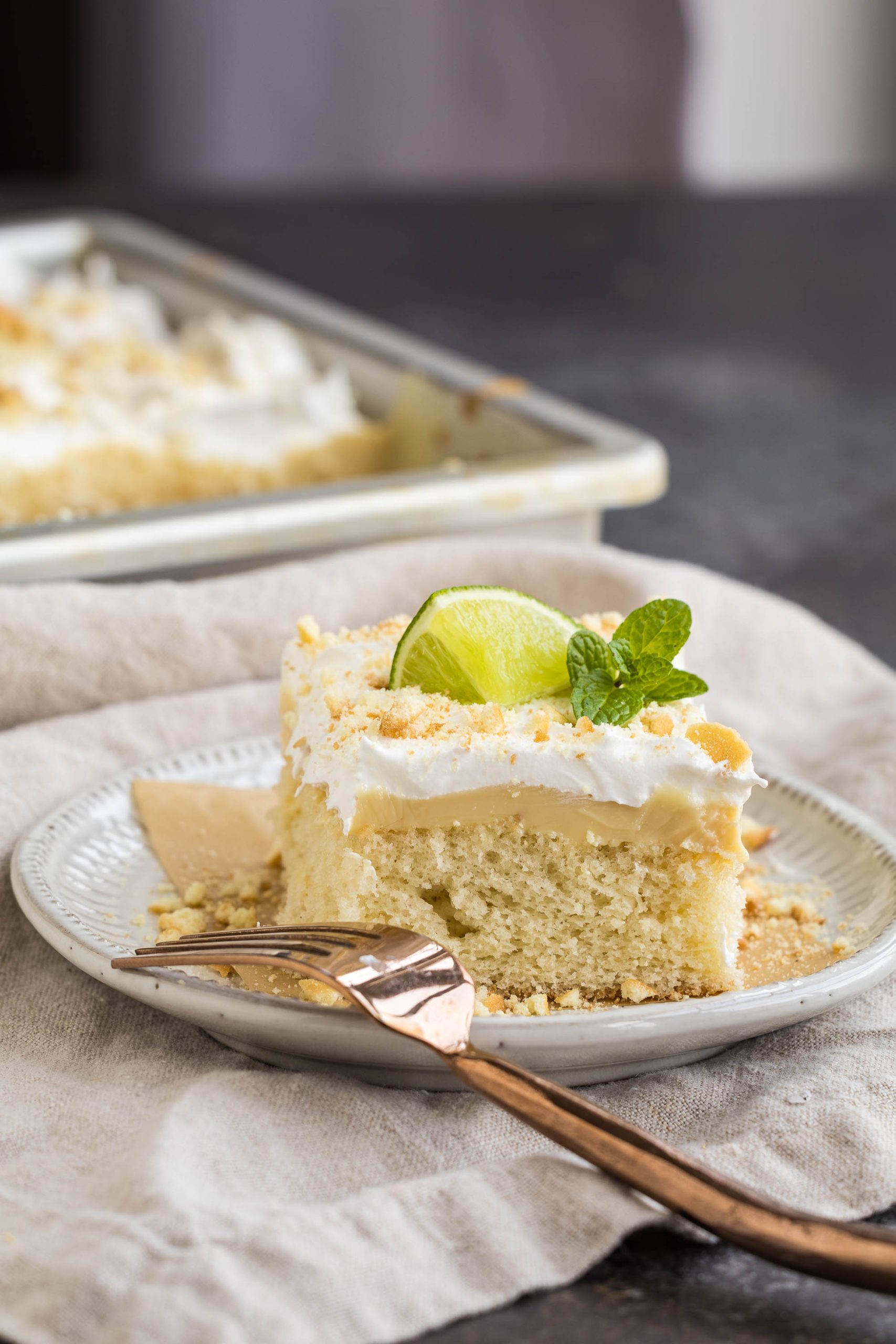 A slice of Key Lime Pie Sheet Cake with frosting, garnished with a lime wedge and mint, sits on a plate with a fork. A pan containing more cake is blurred in the background.