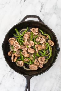 Sliced mushrooms and green bell peppers cooking in a black cast iron skillet on a marble countertop.