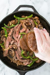 Hand pouring sauce over sliced steak, mushrooms, and green bell peppers in a cast iron skillet.