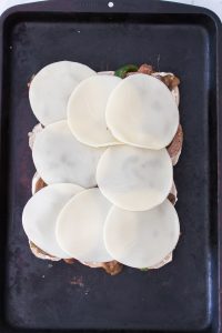 Sliced provolone cheese layered on a sandwich with meat and vegetables on a baking tray.