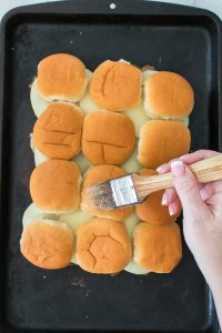 A hand uses a brush to apply butter on top of a batch of slider buns arranged in rows on a dark baking tray.