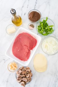 Ingredients for a steak dish are arranged: raw beef, sliced green peppers, onions, mushrooms, butter, olive oil, garlic, pepper, and cheese slices on a marble surface.