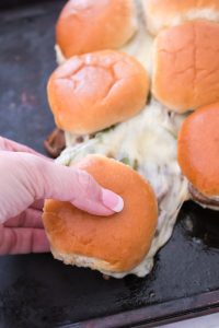 A hand grabbing a slider sandwich with melted cheese from a batch on a baking sheet.