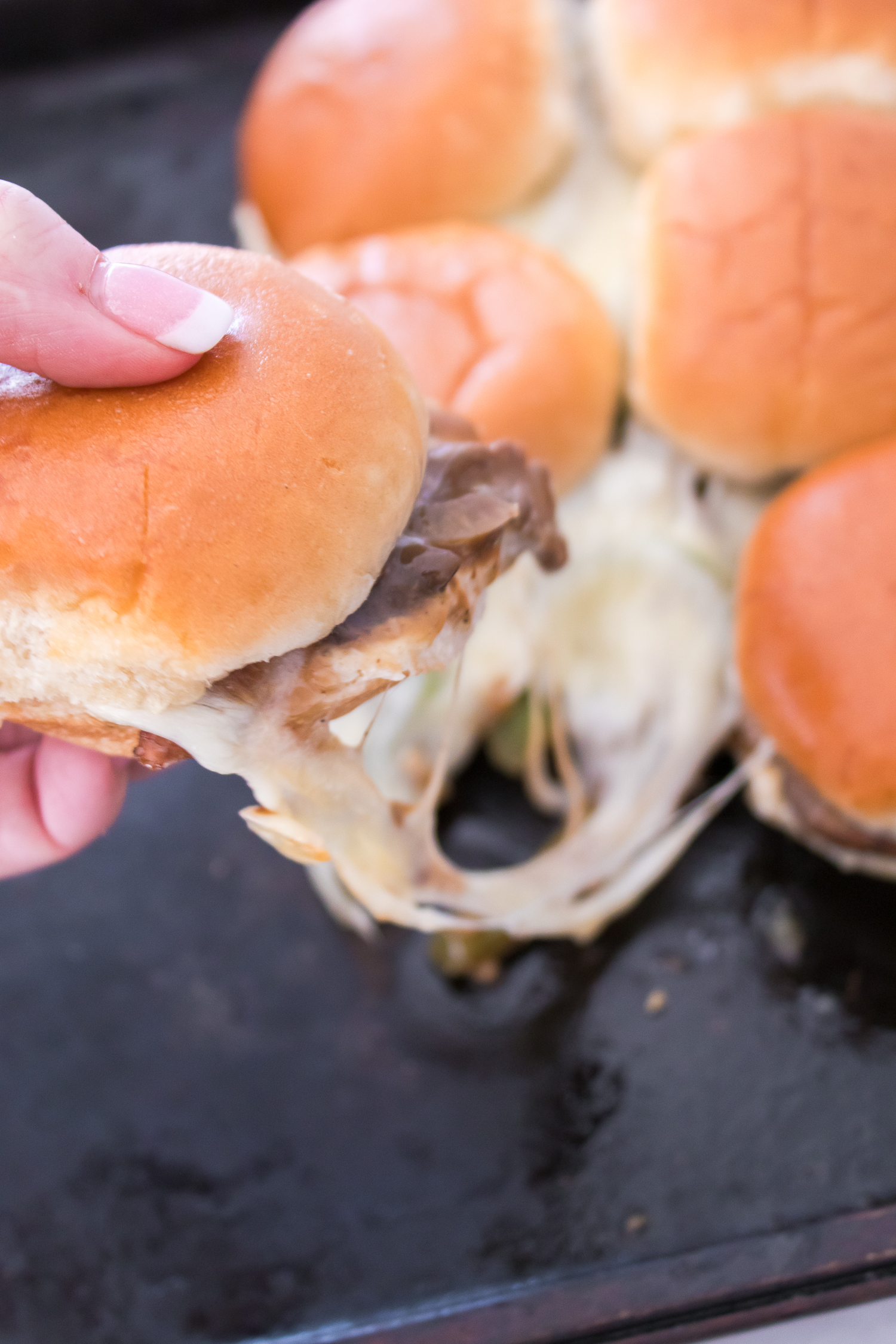 A person lifts a cheesy beef slider from a tray, revealing melted cheese stretched between sliders.