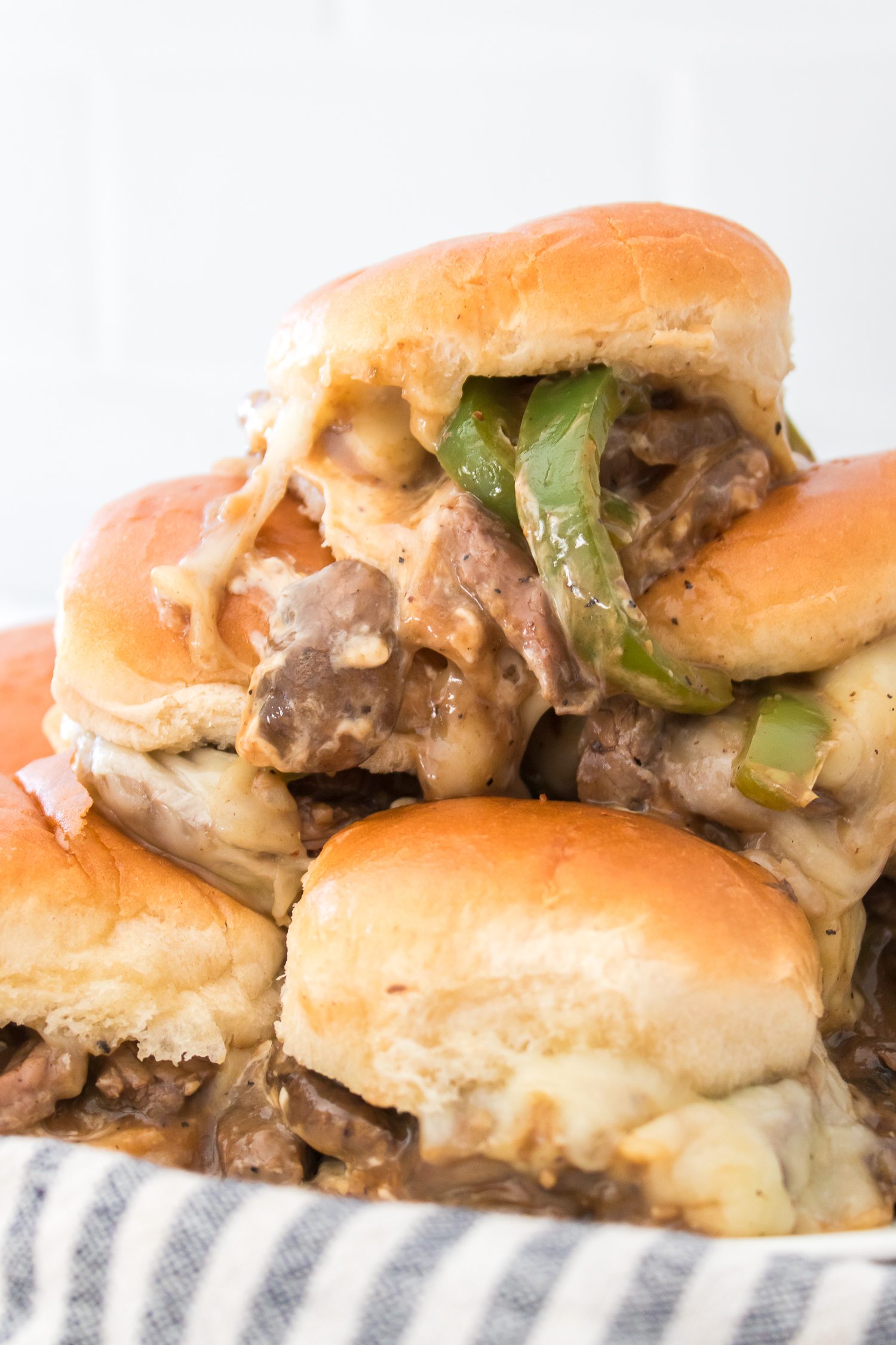 A stack of cheesy Philly Cheesecake Sliders filled with sliced beef and green peppers on a striped cloth.