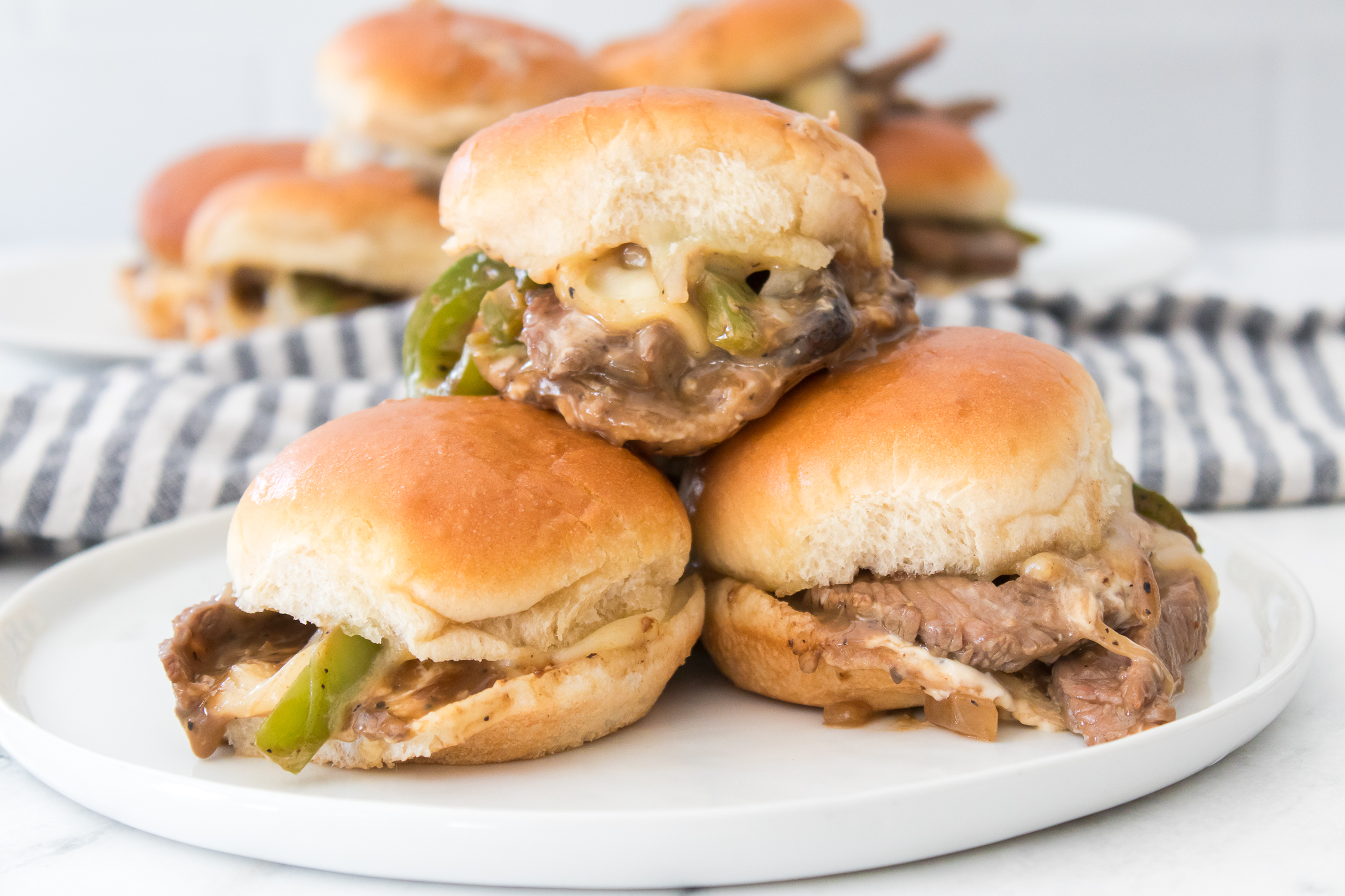 Three Philly Cheesecake Sliders with beef, cheese, and green peppers are stacked on a white plate, with more sliders in the background.