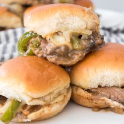 Three sliders with beef, onions, peppers, and cheese on buns are stacked on a white plate. A striped cloth is in the background.