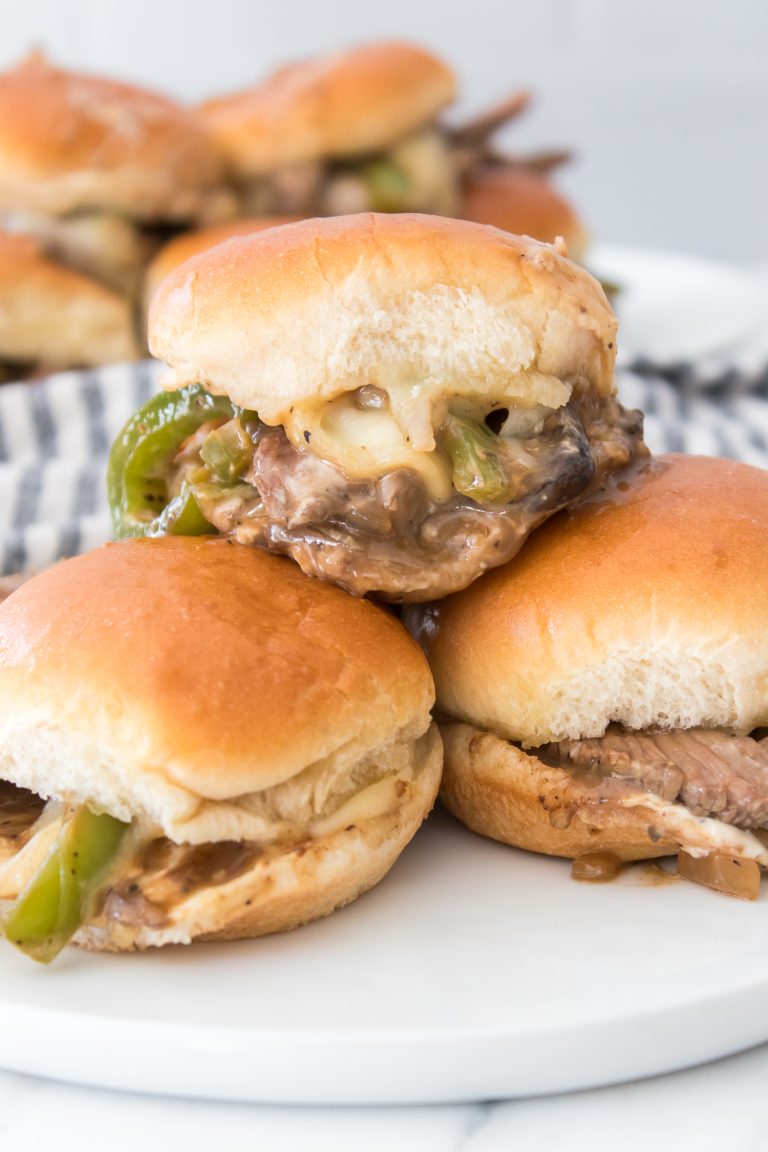 Three sliders with beef, onions, peppers, and cheese on buns are stacked on a white plate. A striped cloth is in the background.