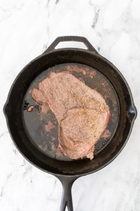 A piece of seasoned steak cooking in a black cast iron skillet on a marble surface.