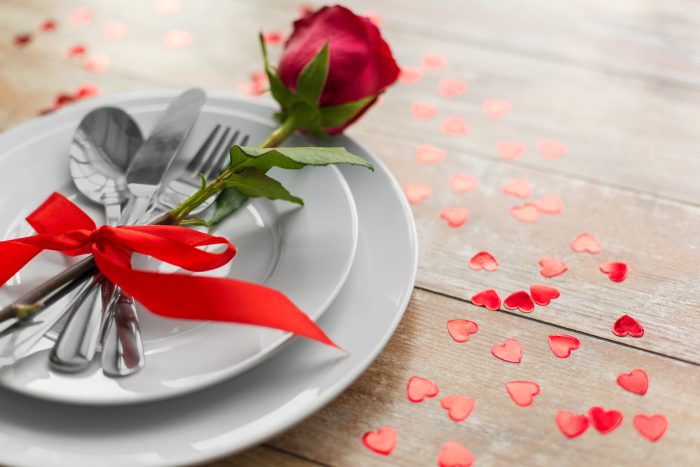 A place setting with a knife, fork, and spoon tied with a red ribbon on white plates, next to a red rose. Red heart-shaped confetti scattered on a wooden table.