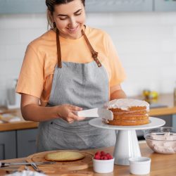 A woman in a kitchen wearing an apron applies frosting to a layer cake on a stand. Nearby are raspberries and a bowl of frosting.