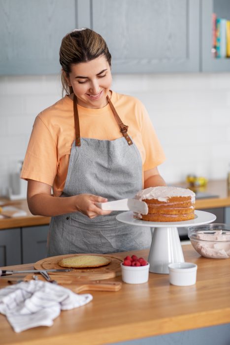 A woman cooking in a kitchen wearing an apron applies frosting to a layer cake on a stand. Nearby are raspberries and a bowl of frosting.