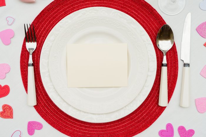 White plate with a blank card on a red placemat. Cutlery is arranged on the sides. Pink and red heart decorations surround the setting.