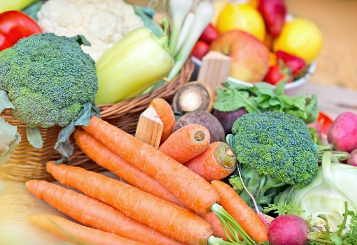 Assorted fresh vegetables and fruits, perfect for recipes to make in March, include carrots, broccoli, peppers, apples, and radishes. They are beautifully arranged on a table near a wicker basket.