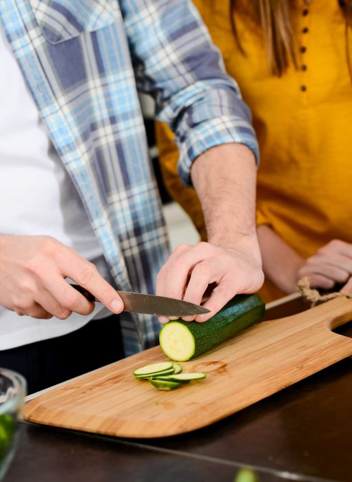 Person slicing a zucchini on a wooden cutting board, with another person standing nearby, wearing a plaid shirt and a yellow top.