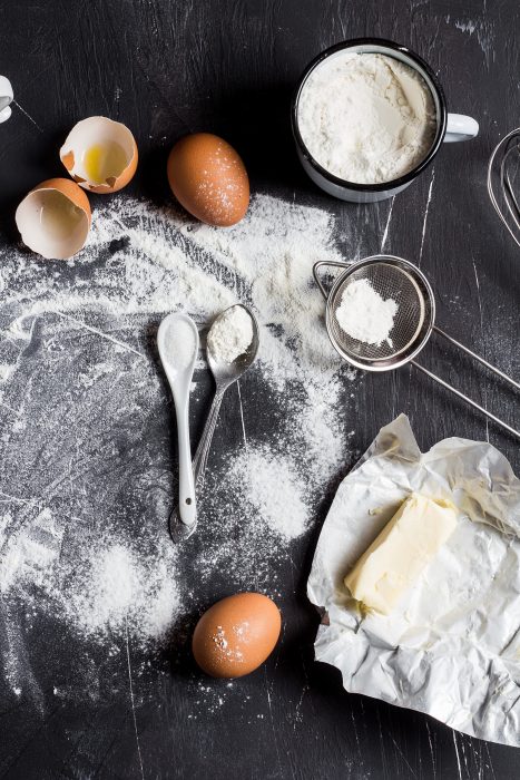 Flour, eggs, and butter on a dark surface with cookie baking tools like a whisk and sifter.