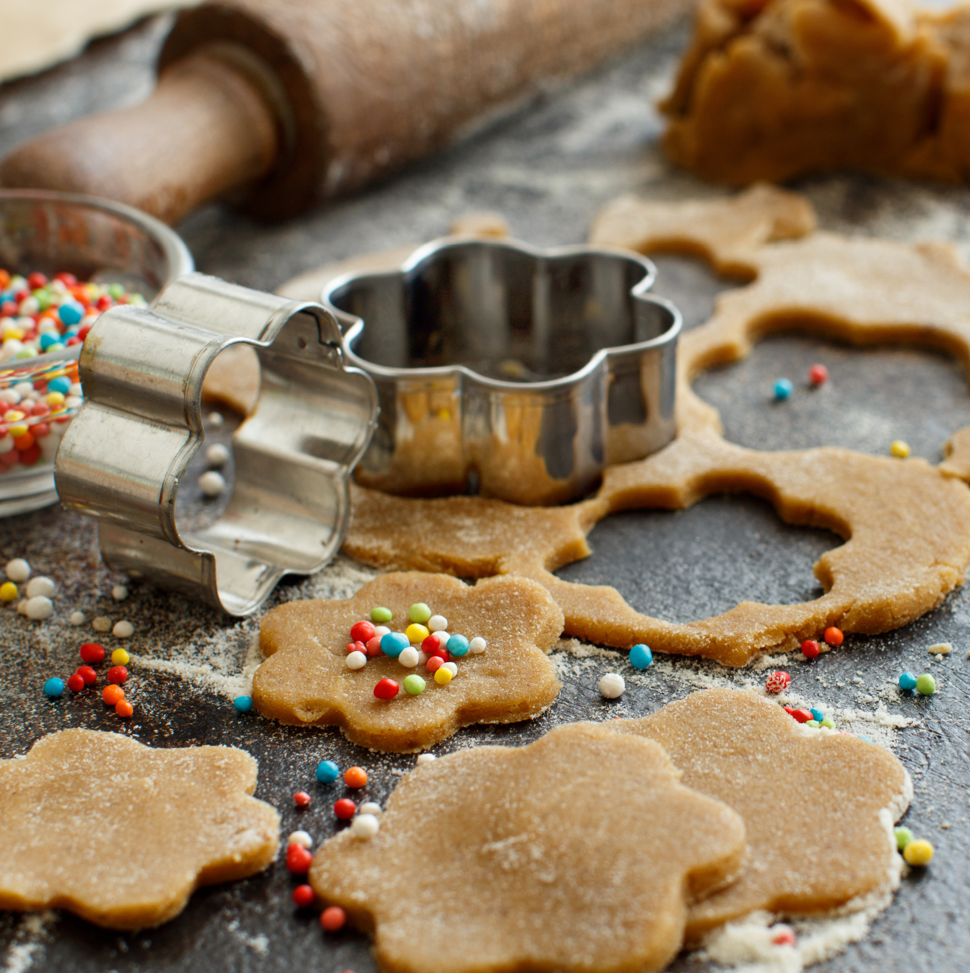 Flower-shaped cookie dough with sprinkles sits alongside an array of cookie baking tools, including metal cutters, on a floured surface.