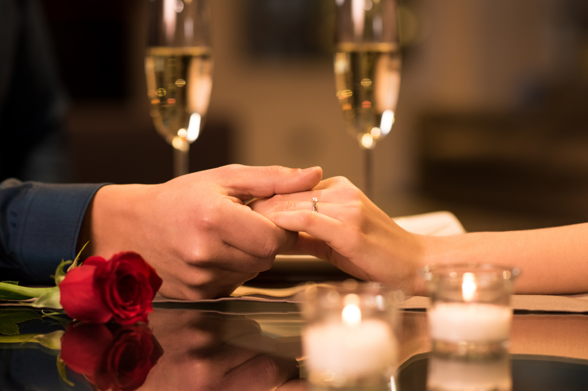 Two people holding hands across a table with lit candles, a rose, and two glasses of champagne present.