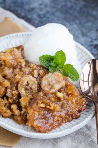 A plate with banana cobbler topped with walnuts and a scoop of vanilla ice cream garnished with a mint sprig. A spoon is placed beside the dessert.