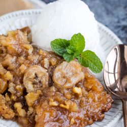 A plate with banana cobbler topped with walnuts and a scoop of vanilla ice cream garnished with a mint sprig. A spoon is placed beside the dessert.