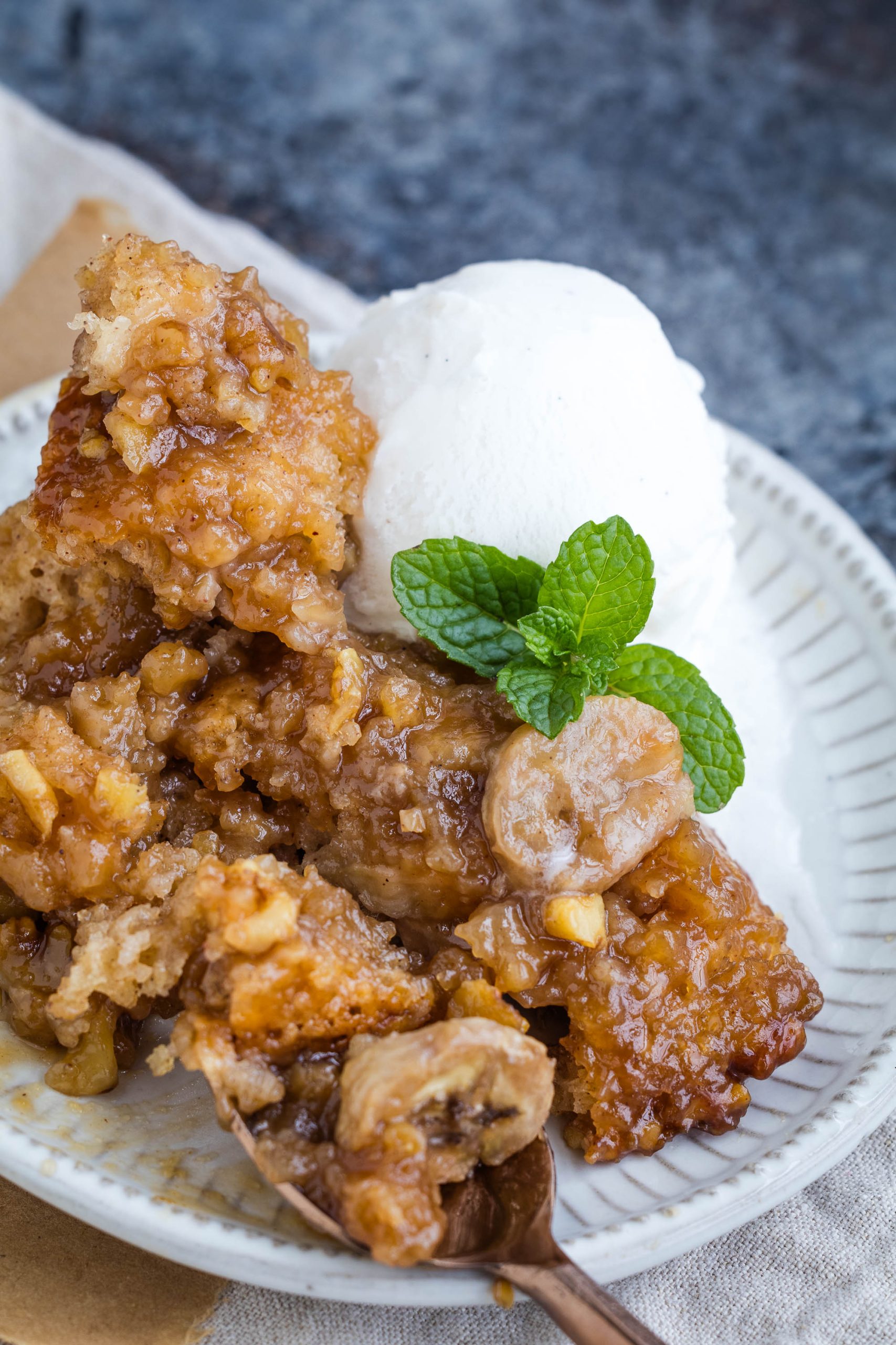 A plate of banana bread pudding topped with ice cream and a mint garnish. A spoon is scooping a portion.