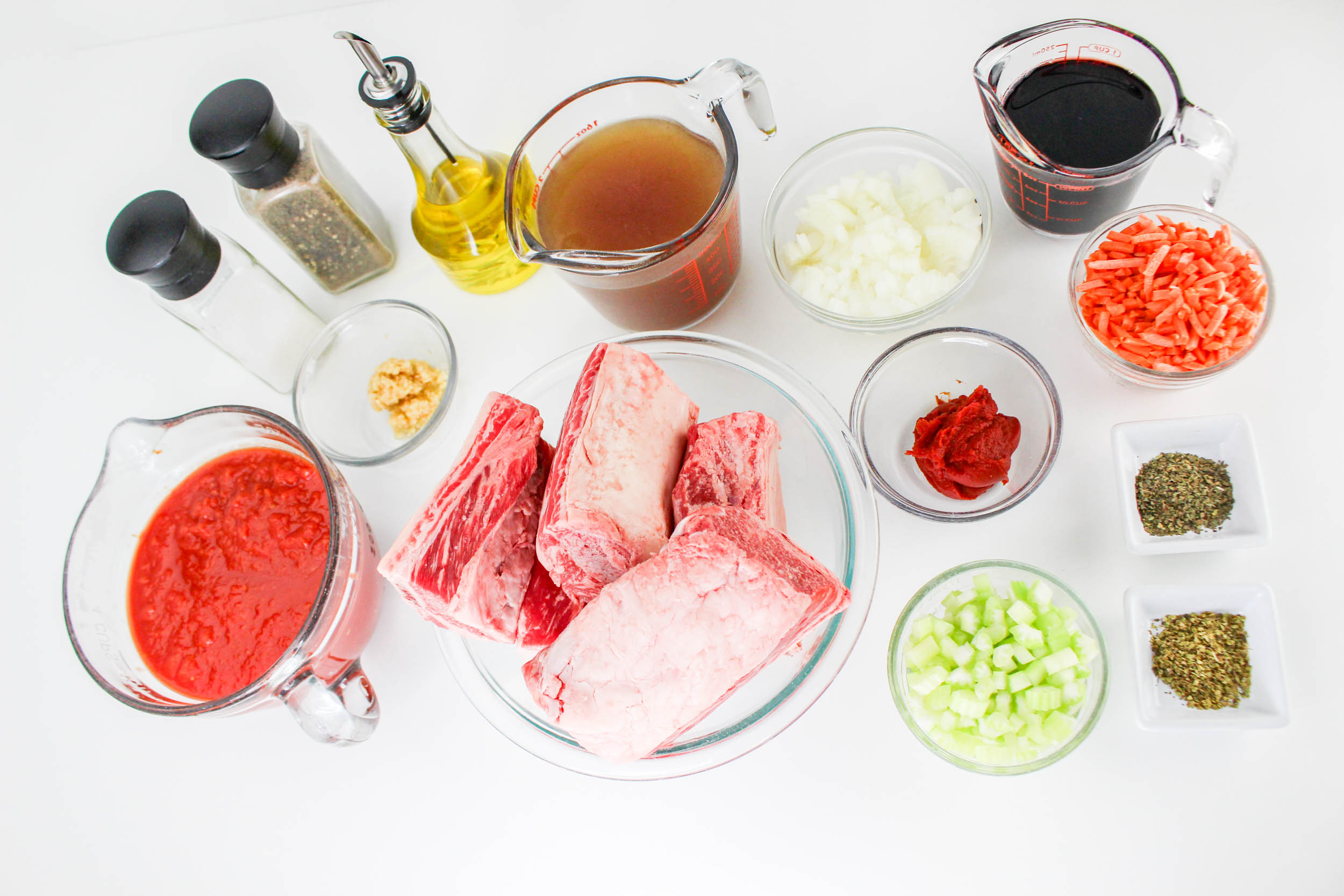 Various cooking ingredients on a white surface include beef short ribs, tomato puree, chopped vegetables, spices, broth, oil, Worcestershire sauce, garlic, salt, and pepper—perfect for creating a rich Bolognese base.