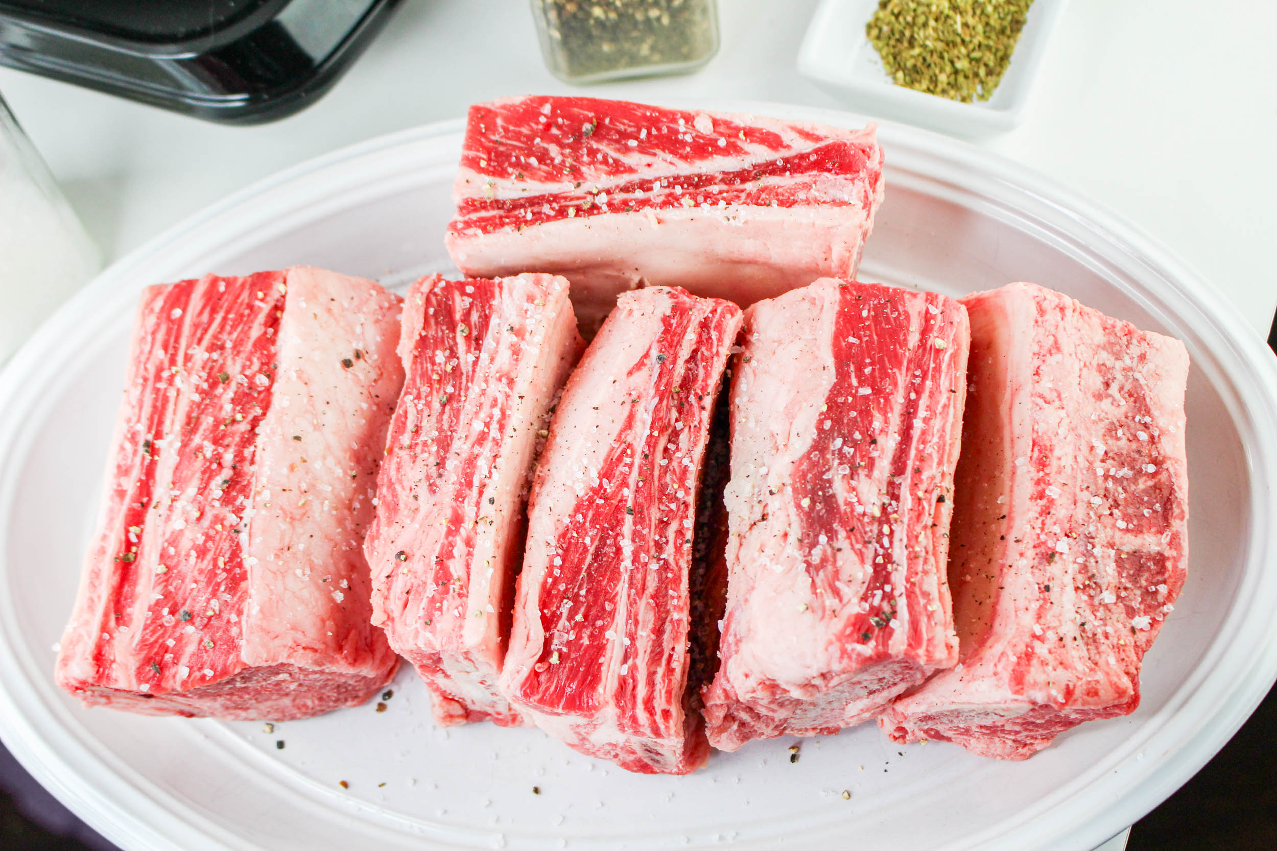 Several raw beef short ribs, seasoned with salt and pepper, are arranged on a white plate, ready to be transformed into a rich Bolognese delight.