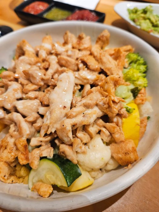 Cantina Del Sol. Bowl of grilled chicken strips over steamed broccoli, cauliflower, and zucchini, topped with sauce. Dipping sauces and guacamole are in the background.