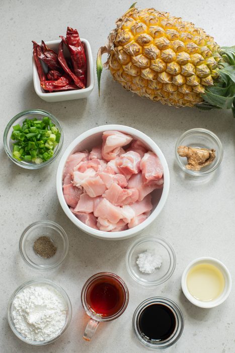 Raw ingredients on a counter: diced chicken in a bowl, whole pineapple, chopped green onions, ginger, black pepper, dried chili peppers reminiscent of stovetop honey pineapple pork. Cornstarch, vinegar, soy sauce, and oil sit ready in small dishes.