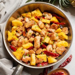 A skillet of cooked chicken with pineapple chunks and red chili peppers, placed on a gray cloth with a wooden spoon and a fresh pineapple nearby.