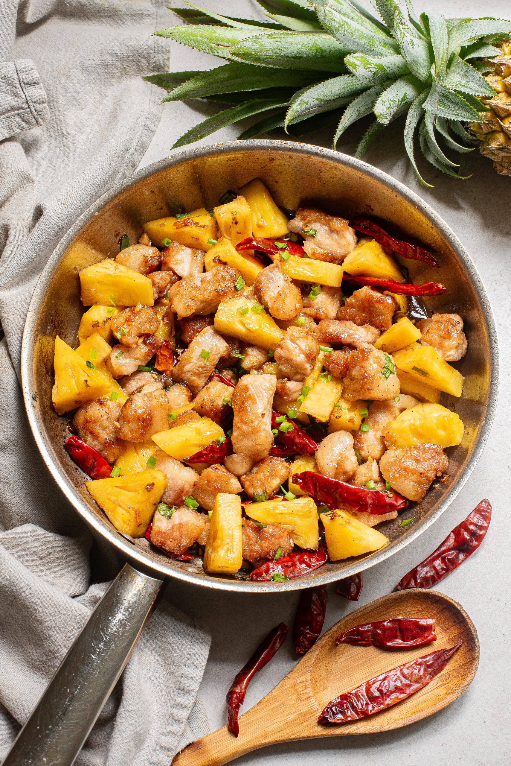 A skillet of cooked chicken with pineapple chunks and red chili peppers, placed on a gray cloth with a wooden spoon and a fresh pineapple nearby.