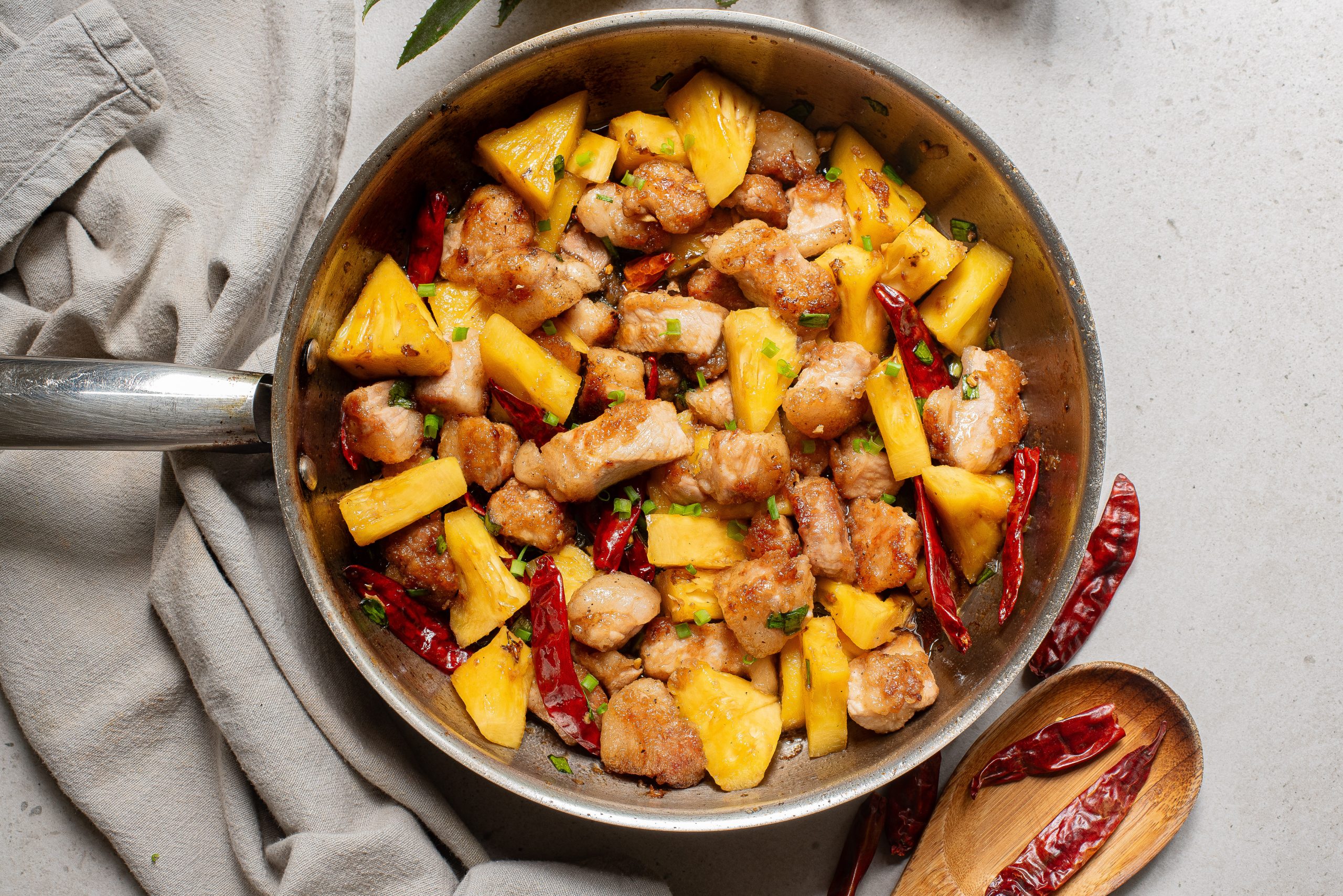 Stir-fried dish with chicken, fresh pineapple chunks, and red chili peppers in a frying pan on a gray surface, with a wooden spoon and scattered chilies nearby.