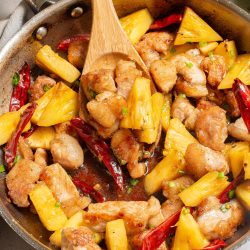 A skillet containing cooked chicken pieces, pineapple chunks, and red chili peppers, with a wooden spatula stirring the mixture.