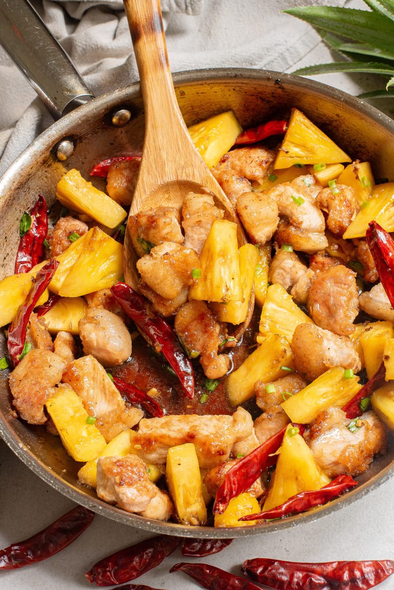 A skillet containing cooked chicken pieces, pineapple chunks, and red chili peppers, with a wooden spatula stirring the mixture.