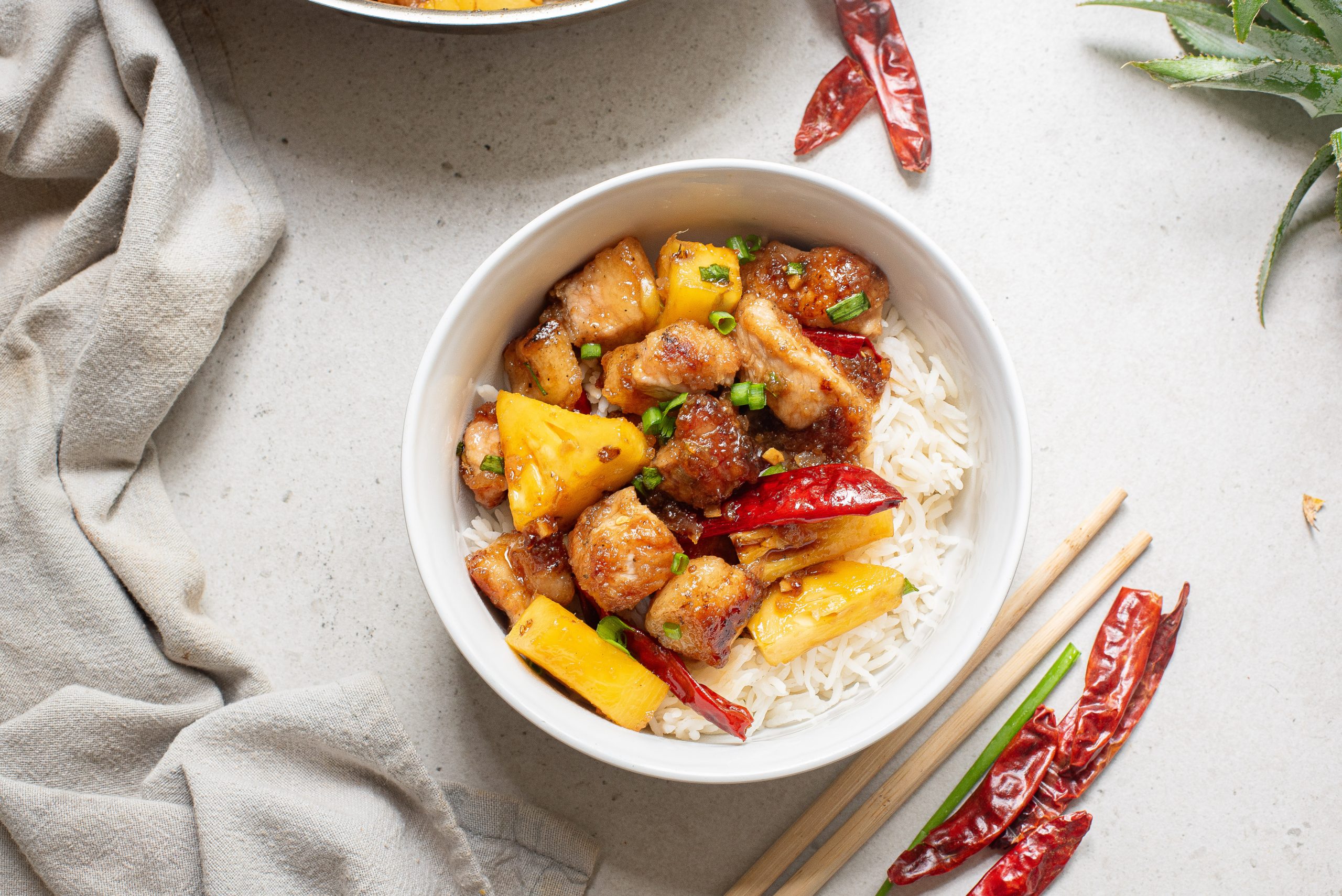 Bowl of rice topped with Stovetop Honey Pineapple Pork. Chopsticks and dried chilies are nearby on a light surface.