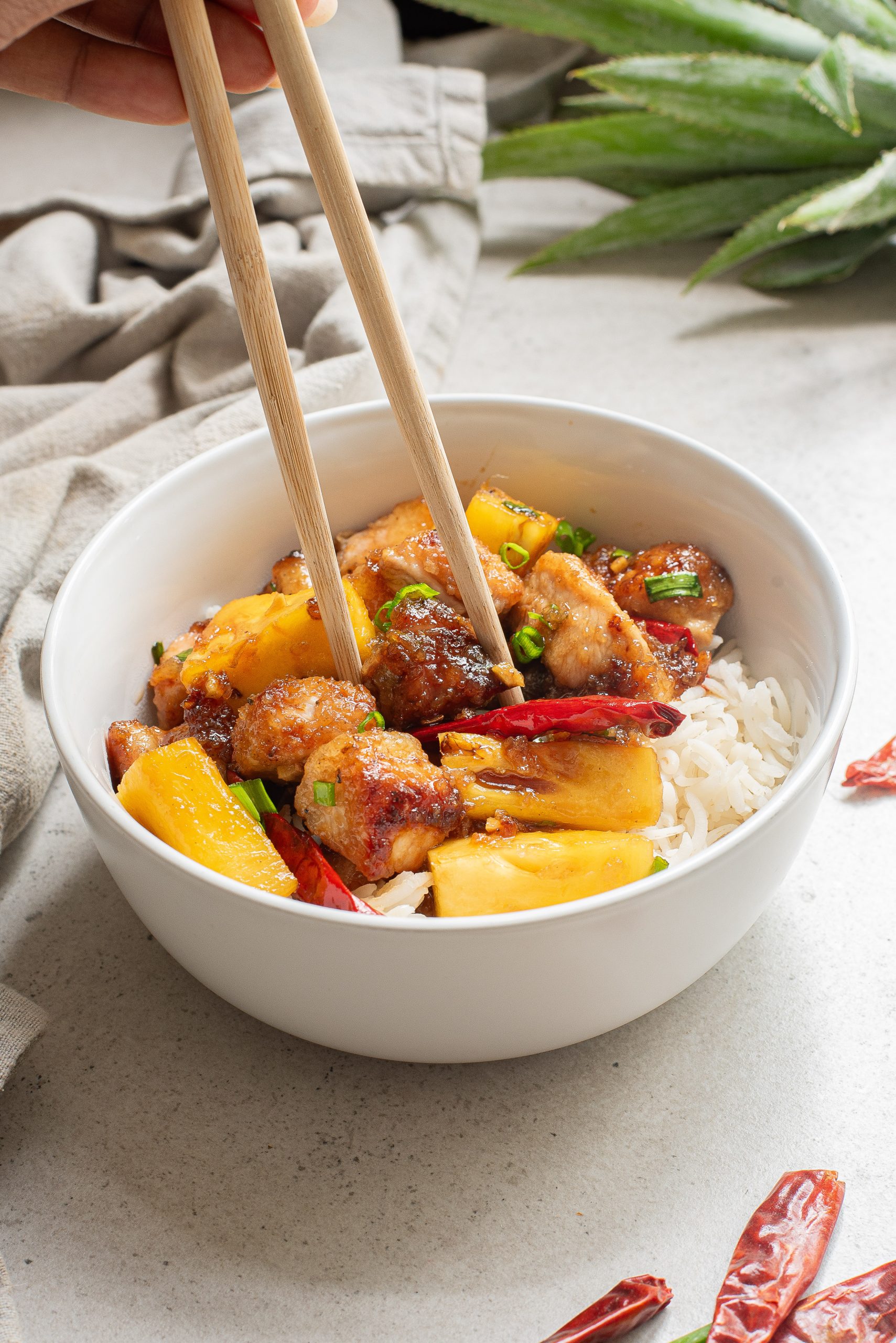 A bowl of rice topped with pineapple chunks, cooked meat pieces, chilies, and scallions. A hand is using chopsticks to pick up the food.