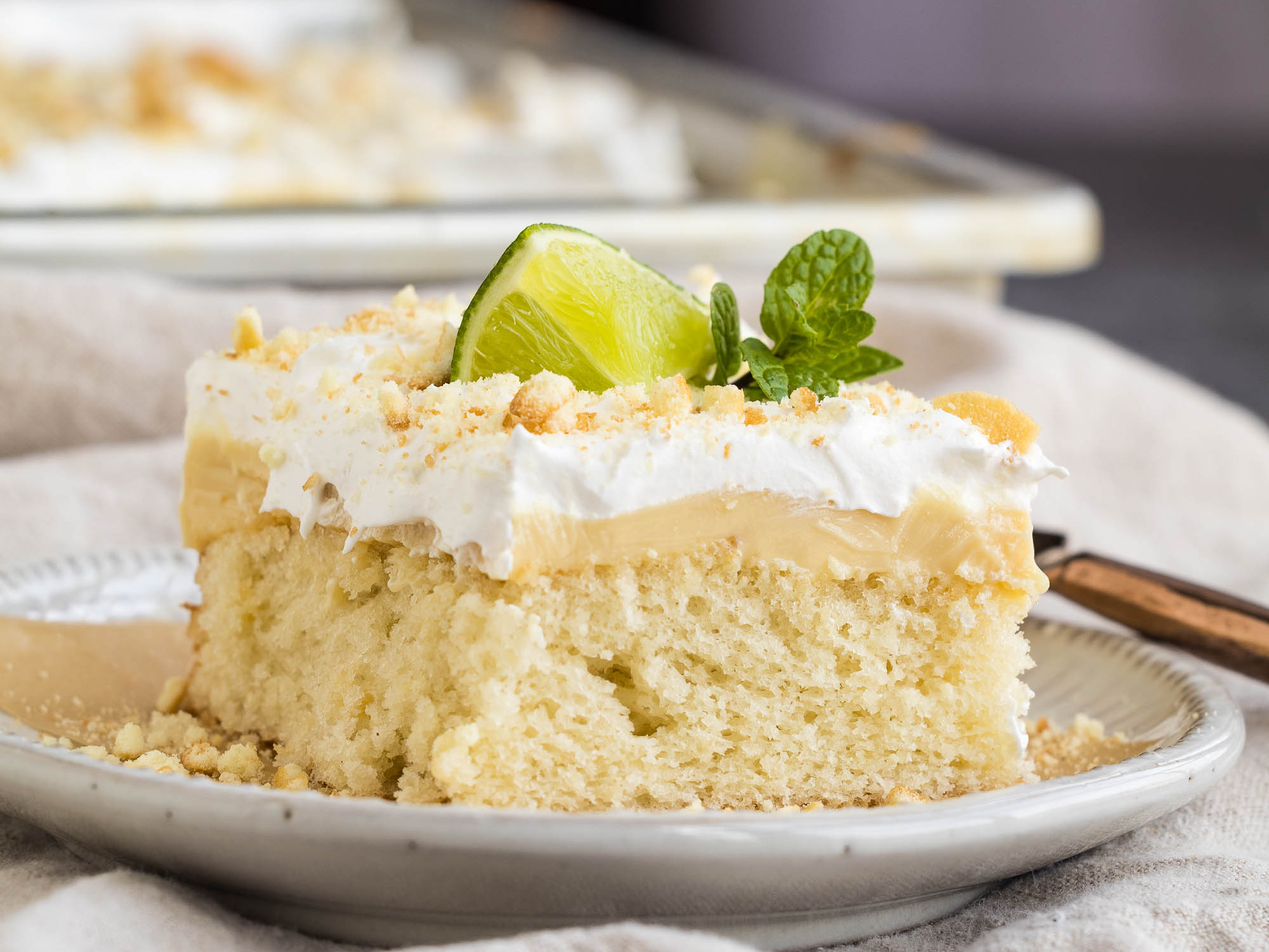 A slice of light cake topped with whipped cream, garnished with a lime wedge and mint, on a white plate.